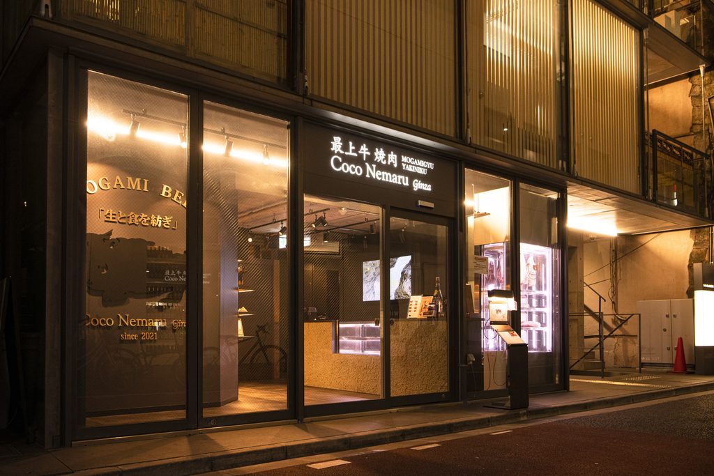Night view of the Coco Nemaru Ginza storefront, a Mogami beef yakiniku restaurant, with a glowing sign above glass doors, warm interior lighting, and a lit dry-aging display case on the right.