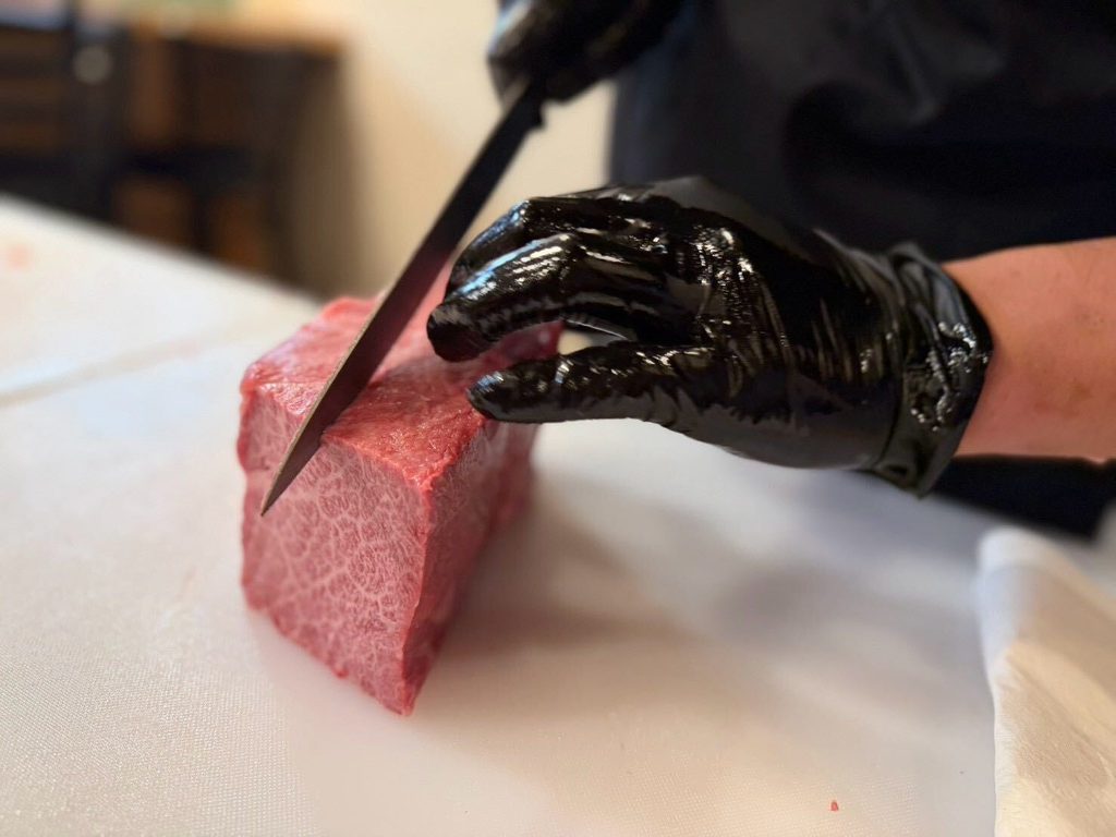 Un carnicero con guantes negros corta un gran trozo de carne de wagyu veteada sobre una tabla de cortar blanca, con la barra de un restaurante japonés visible al fondo.