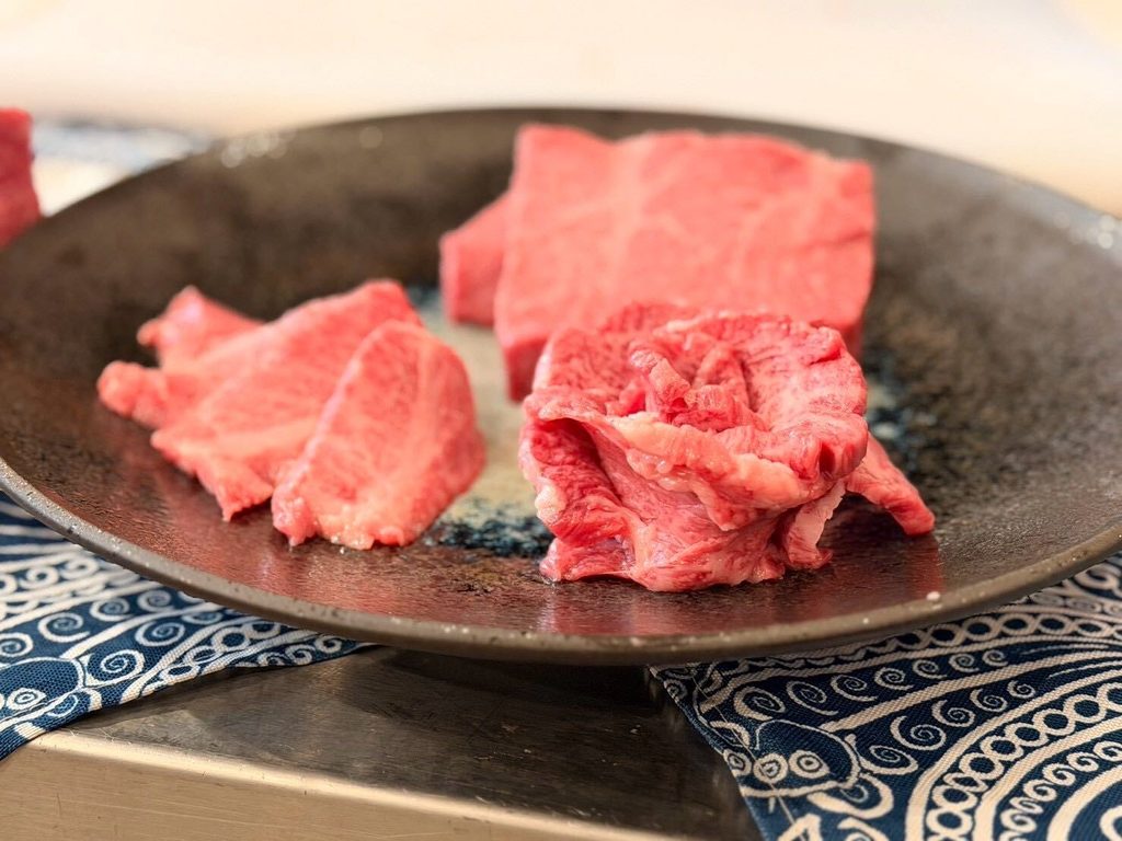 Finas lonchas de carne cruda de wagyu veteada, dispuestas sobre un plato de cerámica oscura colocado sobre un mantel con motivos azules.