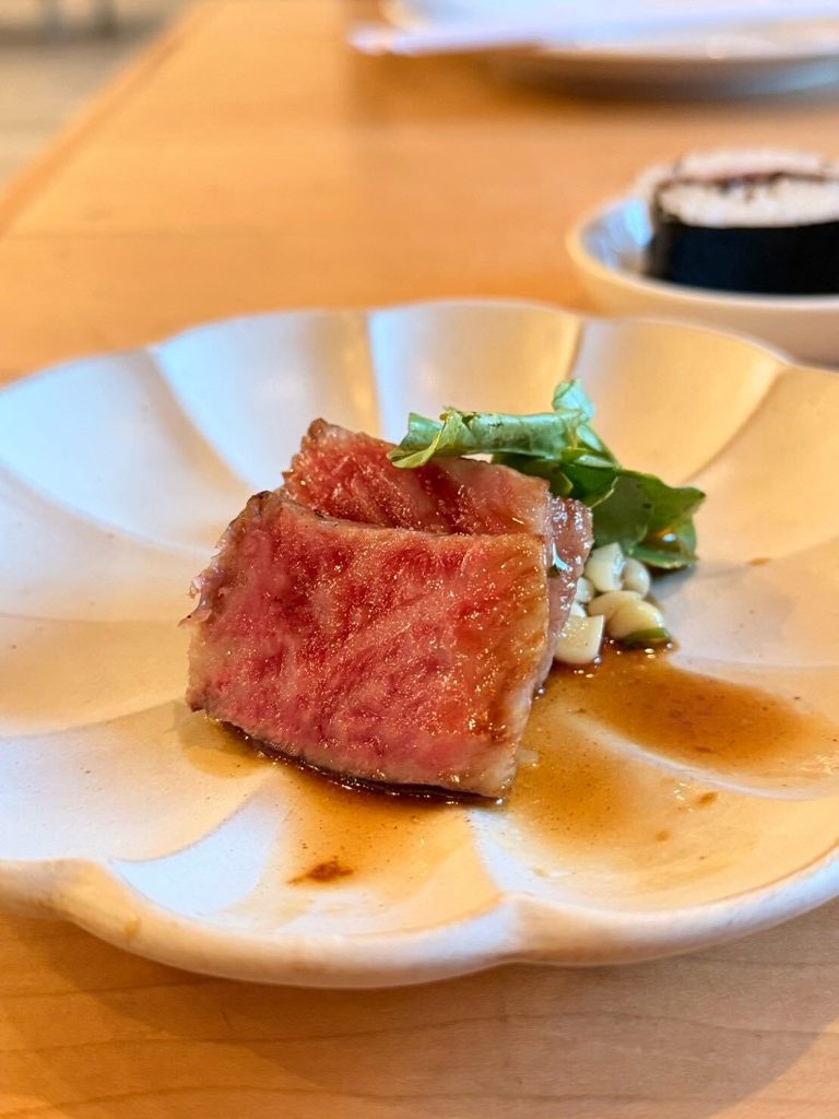 Un cubo de ternera wagyu veteada, sellado a la plancha, servido en un plato blanco festoneado con verduras, chips de ajo y una salsa sabrosa, con un plato de arroz al fondo.