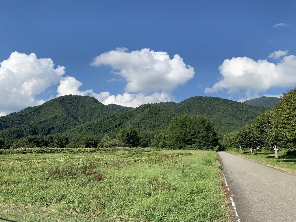 Green mountains under a bright blue sky with large white clouds, above a grassy meadow beside a quiet paved road in Yamagata.