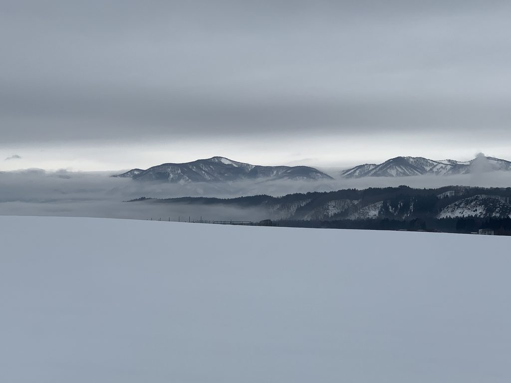 Alt text: Wide snow-covered field under an overcast sky, with mist drifting between snowy mountains in the distance.