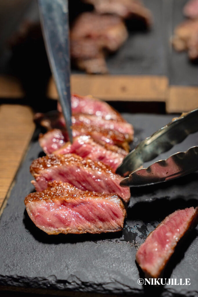 Sliced seared Arita wagyu steak on a slate board, showing a caramelized crust and a juicy pink center, with tongs and a knife.