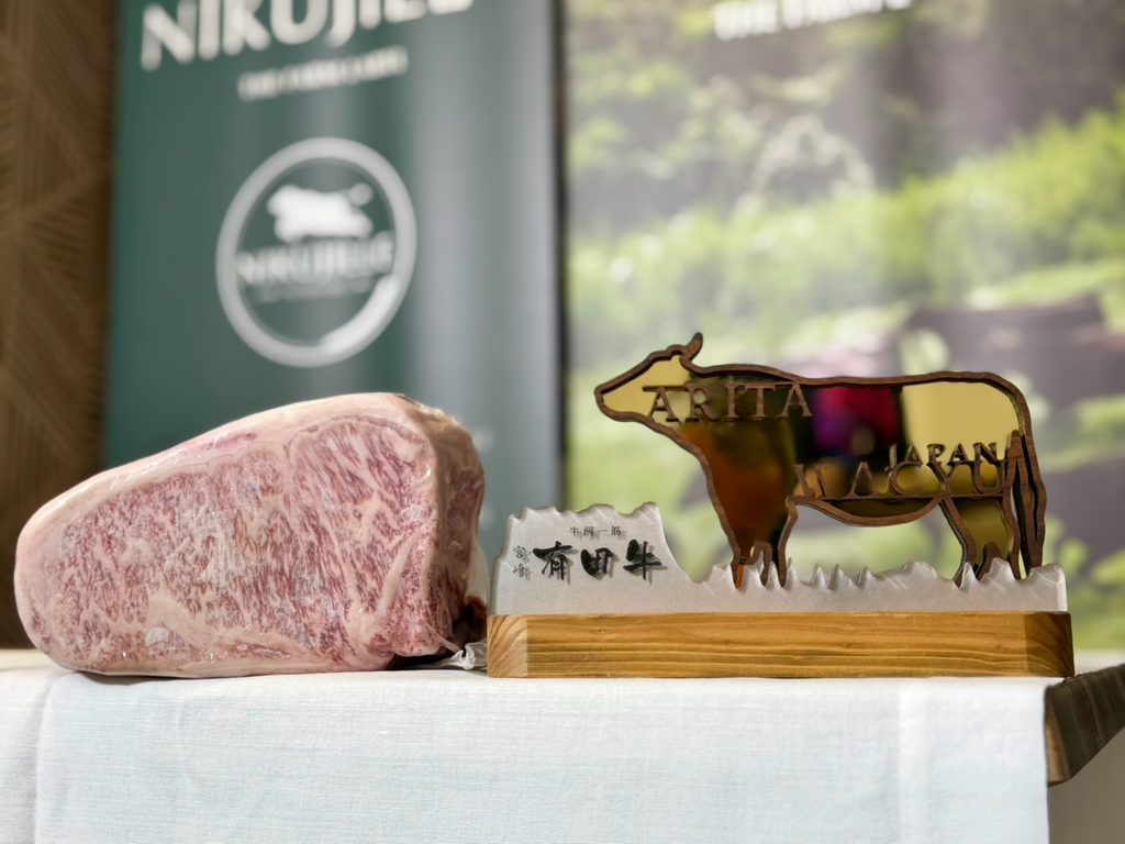 A raw, heavily marbled wagyu striploin on a white tablecloth next to a wooden Arita Wagyu cow-shaped sign with Japanese lettering. A Nikujille banner is blurred in the background.