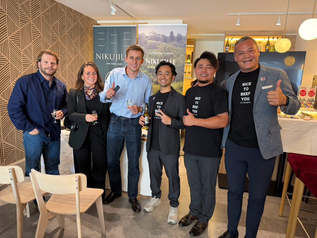 Six people pose together giving thumbs up, some holding drinks. Three of them wear black “Nice to Beef You” Nikujille t-shirts, and the Nikujille banners stand behind them.