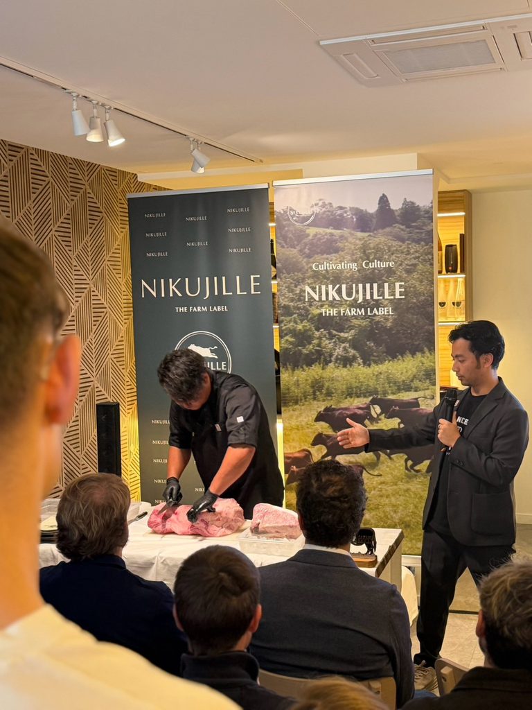 A chef in black gloves breaks down a large piece of raw wagyu on a table while a presenter with a microphone gestures toward him. Guests watch from the foreground in front of Nikujille banners.