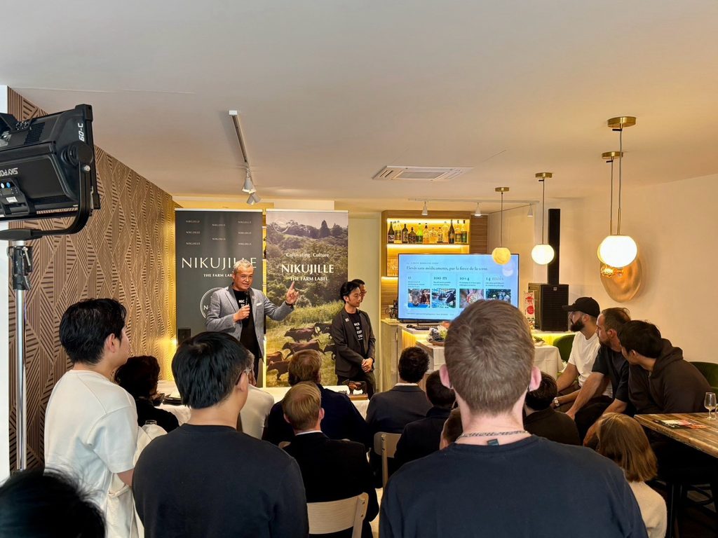 A speaker with a microphone addresses a seated and standing audience in a restaurant. A screen behind him shows a French-language slide about the cattle, with Nikujille banners and a studio light visible.