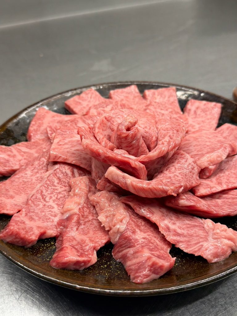Thin slices of raw marbled wagyu arranged in a rose shape on a dark ceramic plate.