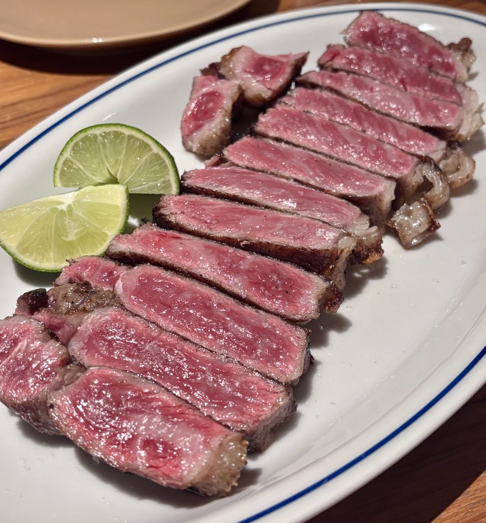 Thick slices of seared wagyu sirloin fanned out on a white oval plate with a blue rim, showing a dark crust and a rosy pink center with fine marbling. Flaky salt is scattered over the meat, and two lime wedges sit at the left edge of the plate.