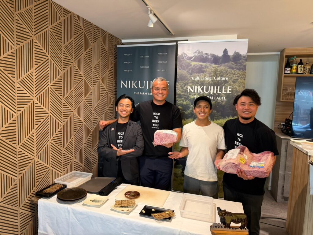 Four smiling men in front of two Nikujille “The Farm Label” banners. Two of them hold raw marbled wagyu cuts, and a table with plates and cutting boards is in front of them.