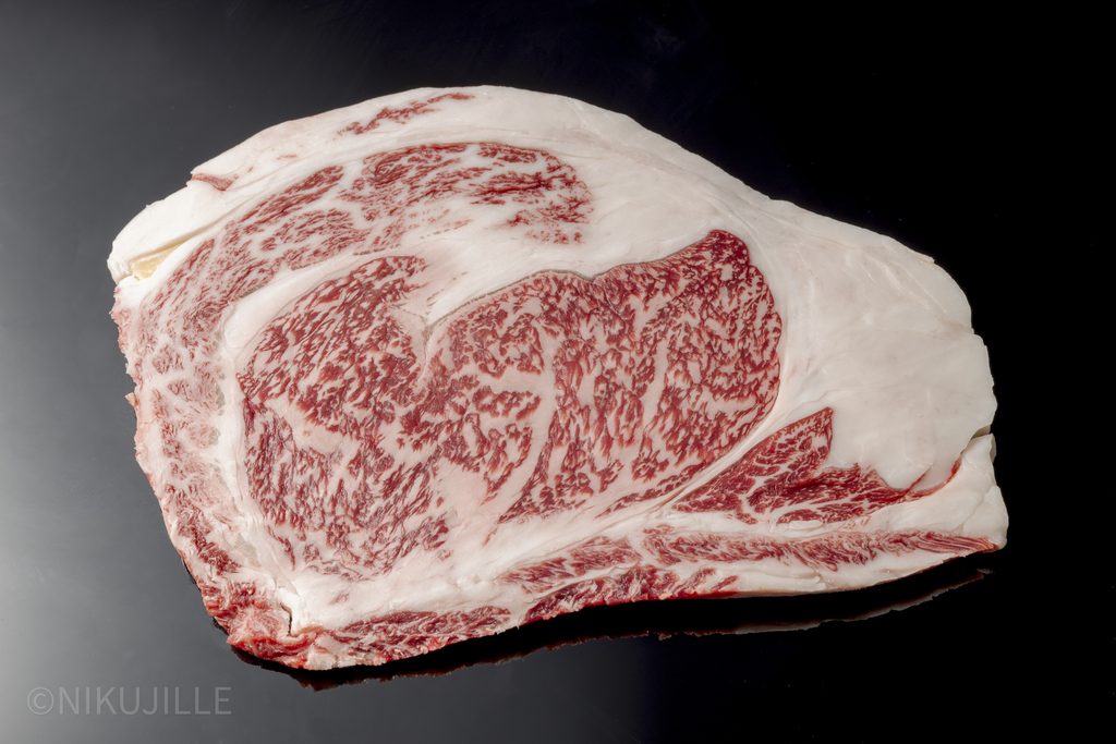 A raw slice of Japanese wagyu ribeye on a black surface, with intricate marbling through the eye of the meat and the rich cap surrounding it.