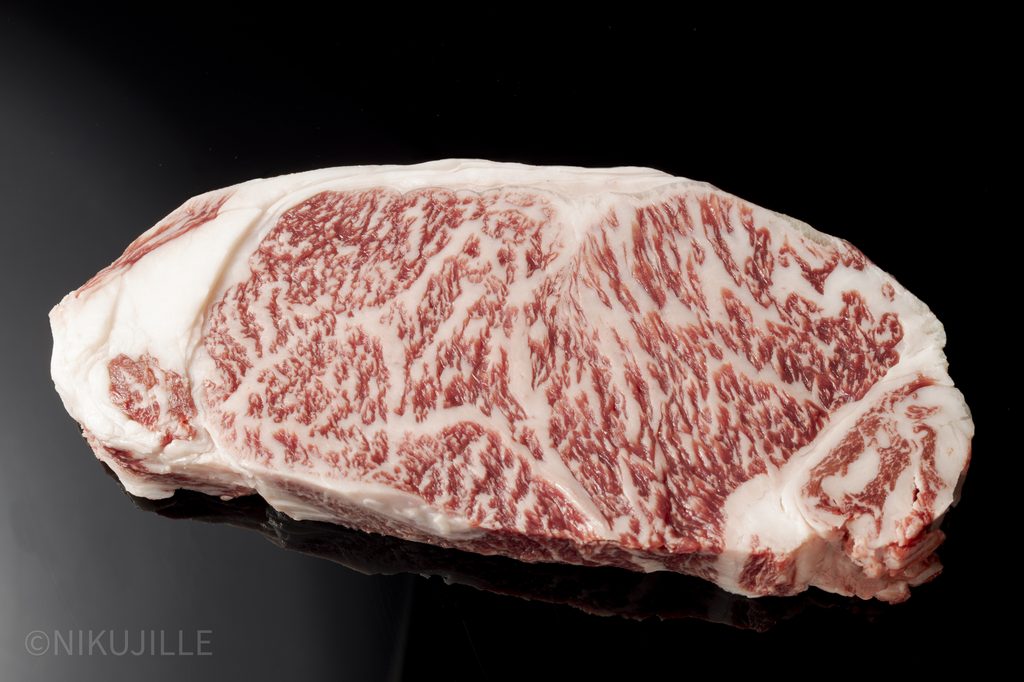 A raw slice of Japanese wagyu striploin on a black surface, showing dense, fine marbling across the cut face with a clean edge of white fat.